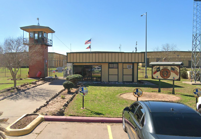 A small, beige building with an American flag on top, next to a watchtower and surrounded by a fence. A parked car is in the foreground.