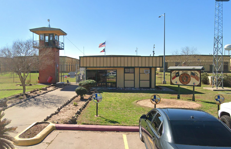 A small, beige building with an American flag on top, next to a watchtower and surrounded by a fence. A parked car is in the foreground.
