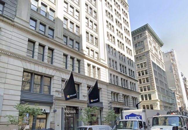 Street view of a Microsoft store with black flags, located in a multi-story, historic-style building with large windows, cars, and a Lowe's truck nearby.