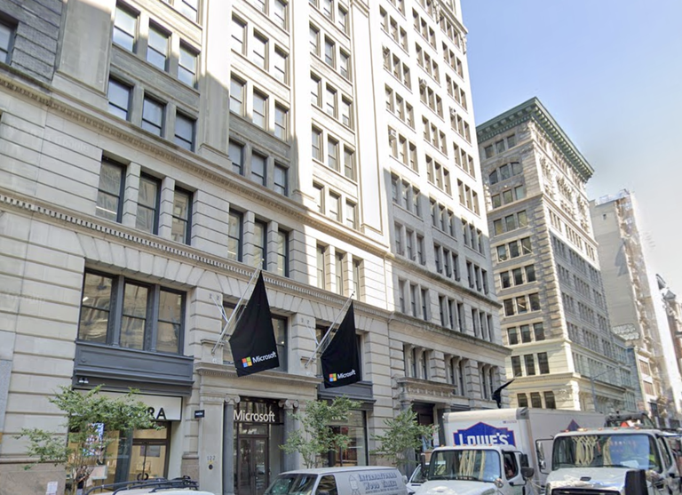 Street view of a Microsoft store with black flags, located in a multi-story, historic-style building with large windows, cars, and a Lowe's truck nearby.