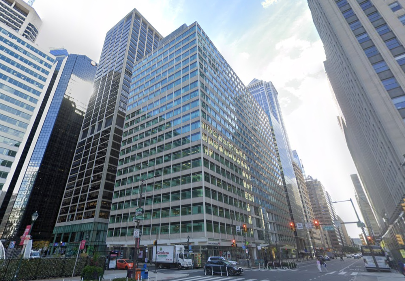 City street view with tall office buildings, traffic lights, a few cars, and pedestrians. The sky is partly cloudy and the area is bustling.