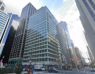 City street view with tall office buildings, traffic lights, a few cars, and pedestrians. The sky is partly cloudy and the area is bustling.