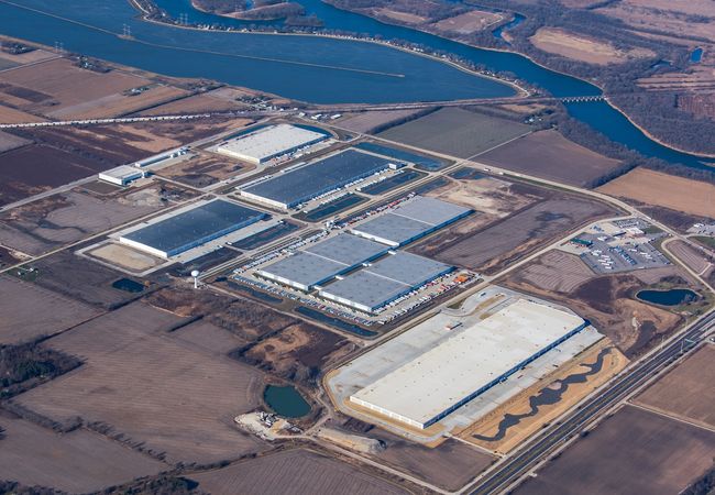 Aerial view of an industrial park with large warehouses surrounded by farmland and a river in the background.
