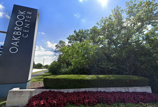 Sign reading "Oakbrook Center" next to lush green trees, a manicured hedge, and vibrant red flowers under a clear blue sky.