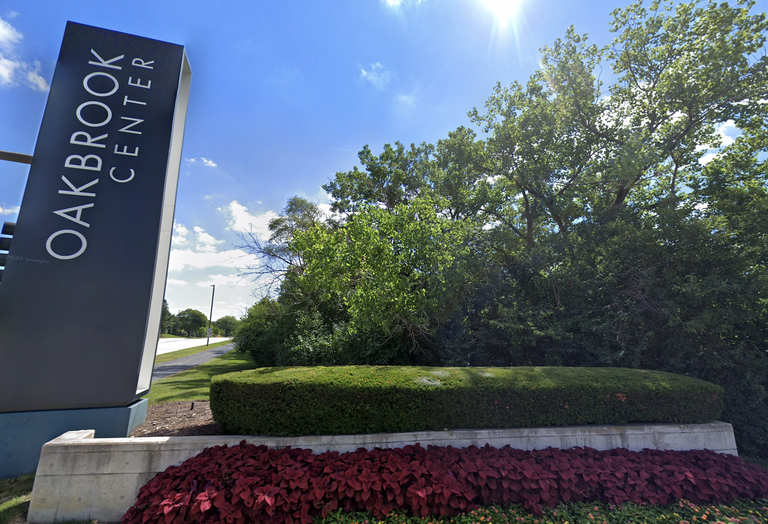 Sign reading "Oakbrook Center" next to lush green trees, a manicured hedge, and vibrant red flowers under a clear blue sky.