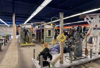 Halloween store display featuring ghoulish figures, a dilapidated cabin marked "Cabin #13," and eerie decorations under fluorescent lights.
