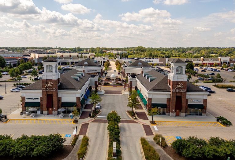 Aerial view of a shopping center with multiple store buildings, large parking areas, and surrounding green space under a partly cloudy sky.