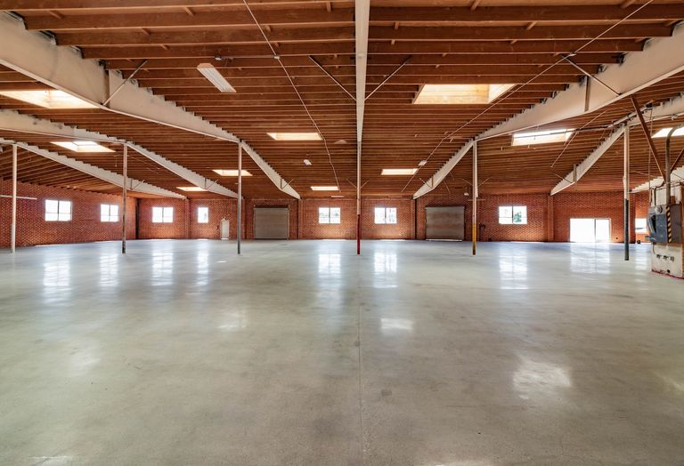 Spacious, empty warehouse interior with polished concrete floor, exposed wooden beams, and red brick walls with windows and skylights.