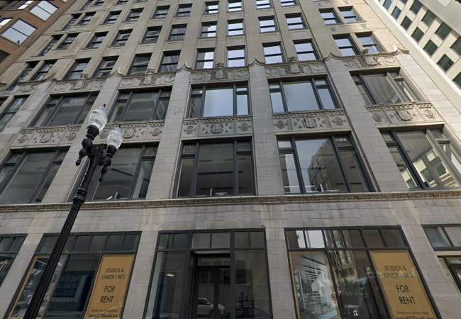 Tall stone building facade with large windows, ornate architectural details, and a "For Rent" sign. A lamp post is visible in the foreground.