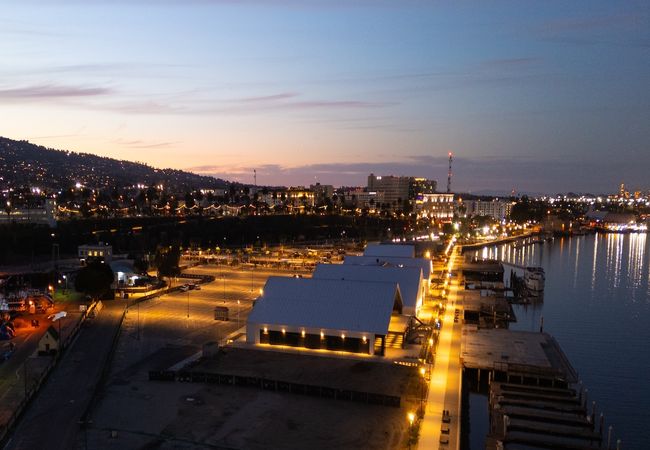 A waterfront cityscape at dusk, featuring illuminated warehouses and a lit pathway along the shoreline.