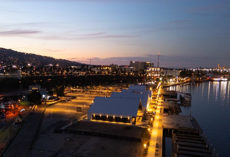 A waterfront cityscape at dusk, featuring illuminated warehouses and a lit pathway along the shoreline.