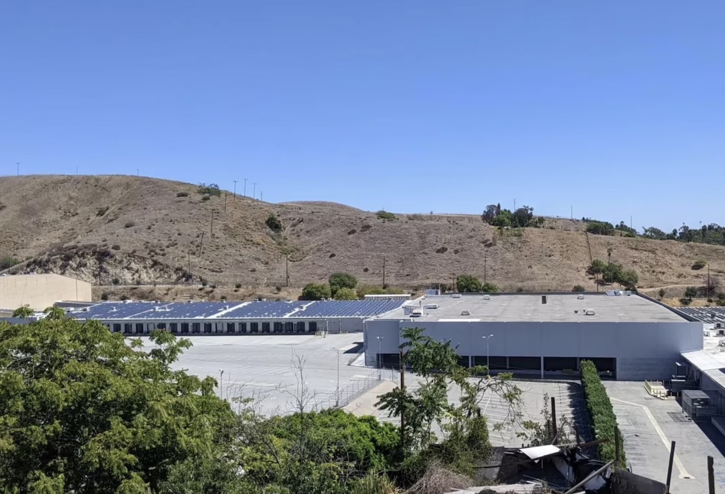 Industrial building with a flat roof and solar panels, set against a backdrop of dry hills and clear blue sky.