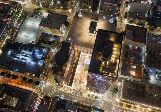 Aerial view of an illuminated city block-sized shopping center and office complex at night, showing tall buildings, streets, and parking lots with bright lights and traffic.