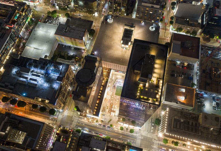 Aerial view of an illuminated city block-sized shopping center and office complex at night, showing tall buildings, streets, and parking lots with bright lights and traffic.