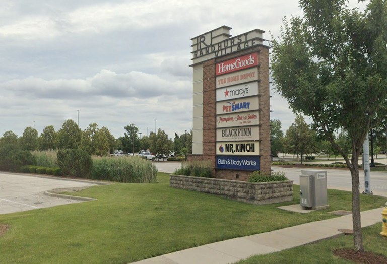 A tall shopping center sign listing stores like HomeGoods, Home Depot, Macy's, and others, set against a grassy area and a cloudy sky.
