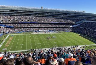 A crowded football stadium with fans watching a game on a sunny day. Players are gathered at the center of the field.