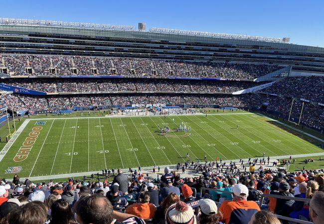 A crowded football stadium with fans watching a game on a sunny day. Players are gathered at the center of the field.