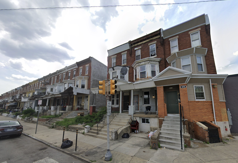 Residential street with a row of brick townhouses, each with steps leading to the entrance, under a partly cloudy sky.