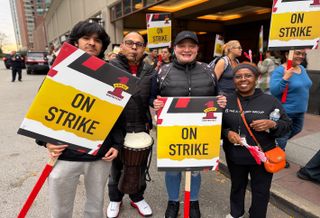 Four people holding "On Strike" signs outside a building, participating in a strike.