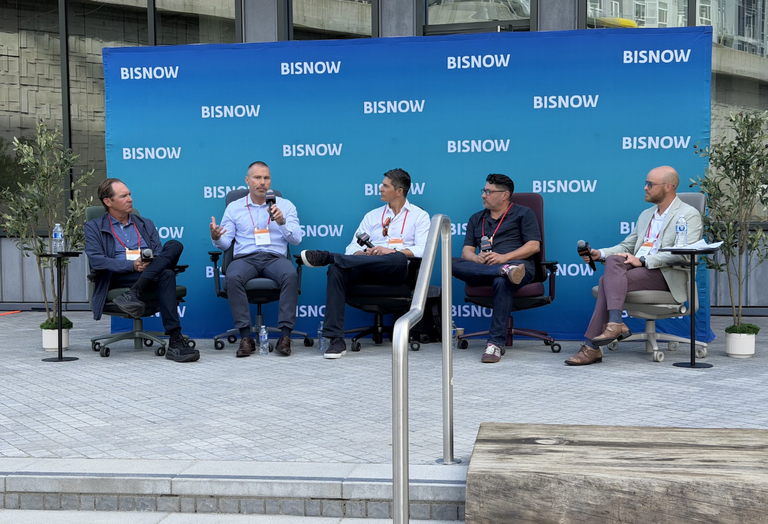 Five people sitting on a panel stage holding microphones, engaged in discussion, with a blue Bisnow backdrop behind them.