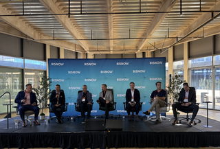 Seven men seated onstage in front of a Bisnow banner, engaged in a panel discussion.
