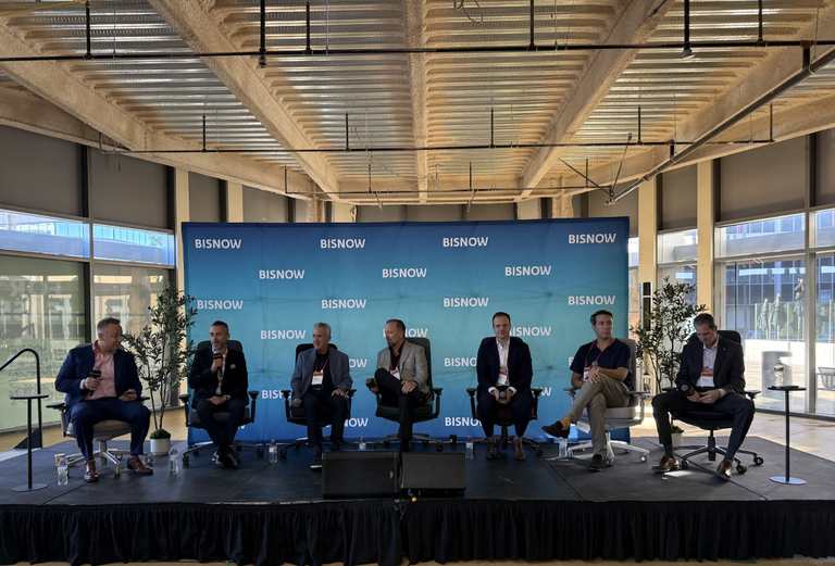 Seven men seated onstage in front of a Bisnow banner, engaged in a panel discussion.