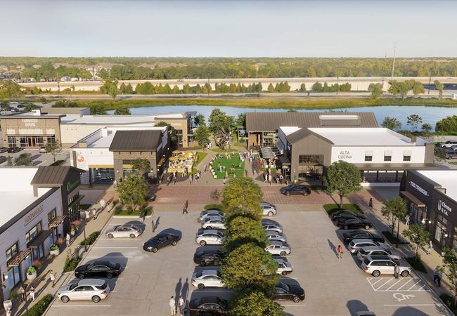 Aerial view of a shopping center with various shops, restaurants, and a central grassy area. Cars are parked in the lot, and people walk around.