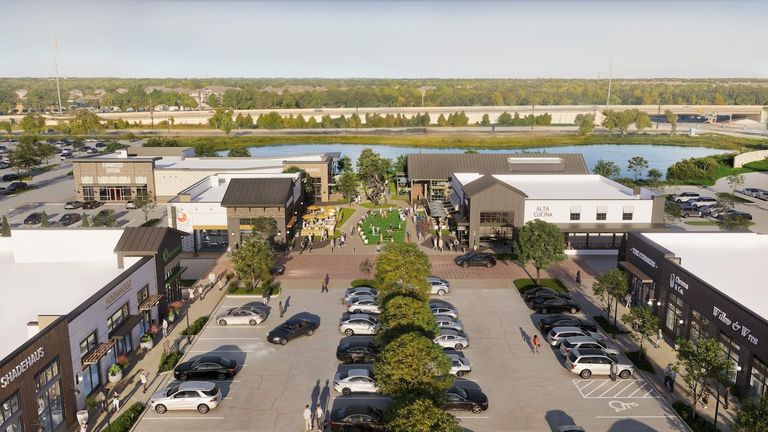 Aerial view of a shopping center with various shops, restaurants, and a central grassy area. Cars are parked in the lot, and people walk around.