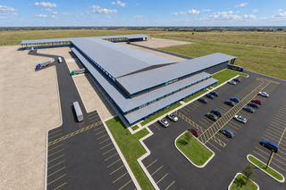 Aerial view of a large industrial warehouse with trucks and trailers parked. Adjacent parking lot has several cars. Surrounded by open grassy fields.