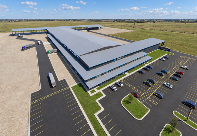 Aerial view of a large industrial warehouse with trucks and trailers parked. Adjacent parking lot has several cars. Surrounded by open grassy fields.