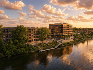 Urban riverside scene at sunset with modern buildings, lush trees, and a bridge in the background, reflecting a warm golden glow on the water.