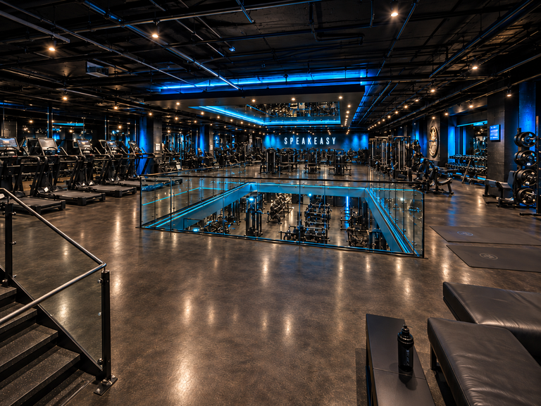 A gym with treadmills, weight machines, mirrors, and blue lighting. A glass partition overlooks a lower level.