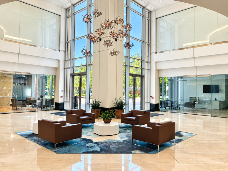 A modern, spacious lobby with high ceilings, large glass windows, brown chairs, a blue rug, plants, and a contemporary chandelier.