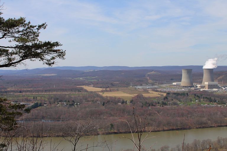 Scenic landscape with a river, fields, and a power plant with two cooling towers emitting steam in the background.