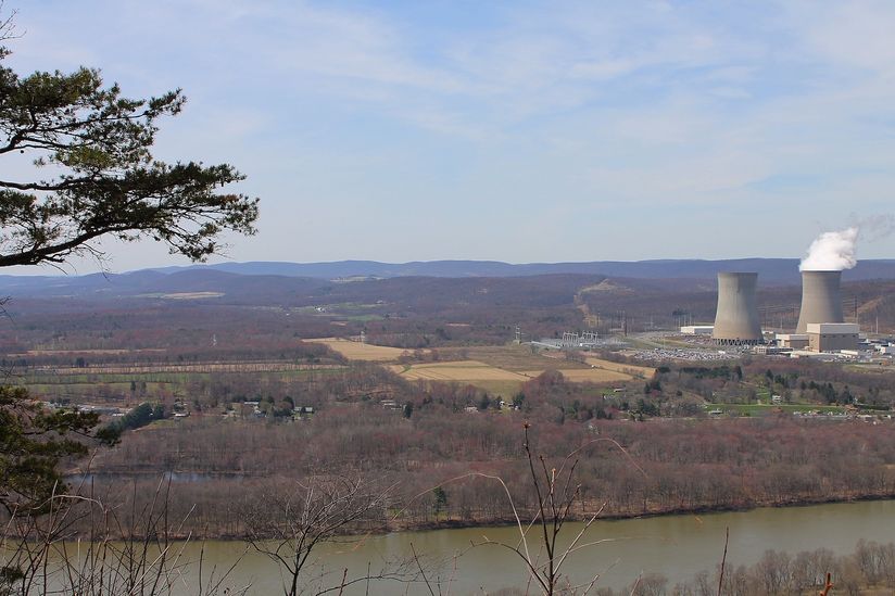 Scenic landscape with a river, fields, and a power plant with two cooling towers emitting steam in the background.