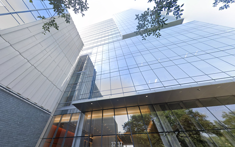 Modern glass skyscraper with reflective windows, surrounded by trees and a clear sky, viewed from below at an upward angle.