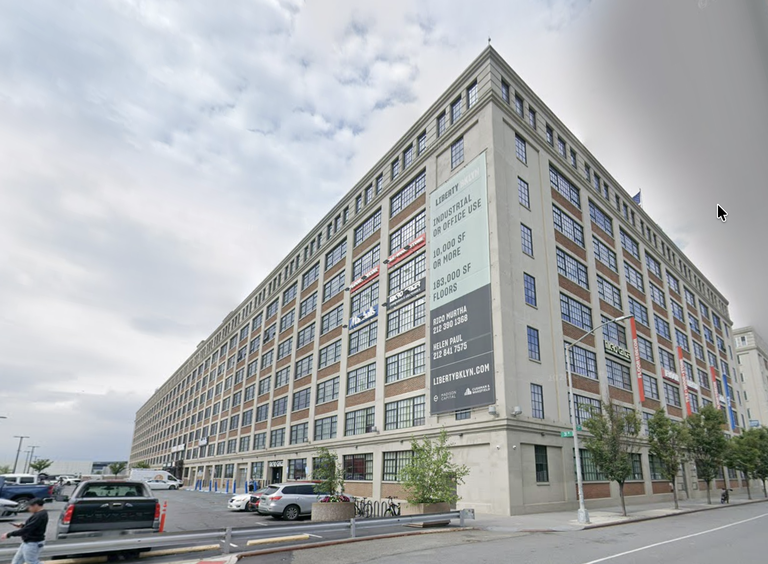 Large multi-story industrial building with a banner for leasing office space, several cars parked in front, and overcast sky.