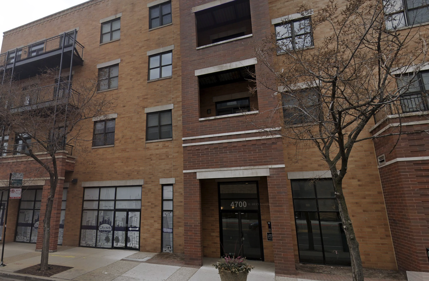 Brick apartment building with multiple floors, windows, and two trees in front on a sidewalk.