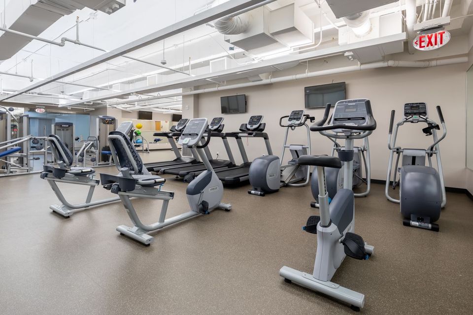 A gym with various exercise equipment, including treadmills, stationary bikes, and ellipticals. The room has a white ceiling and beige walls.