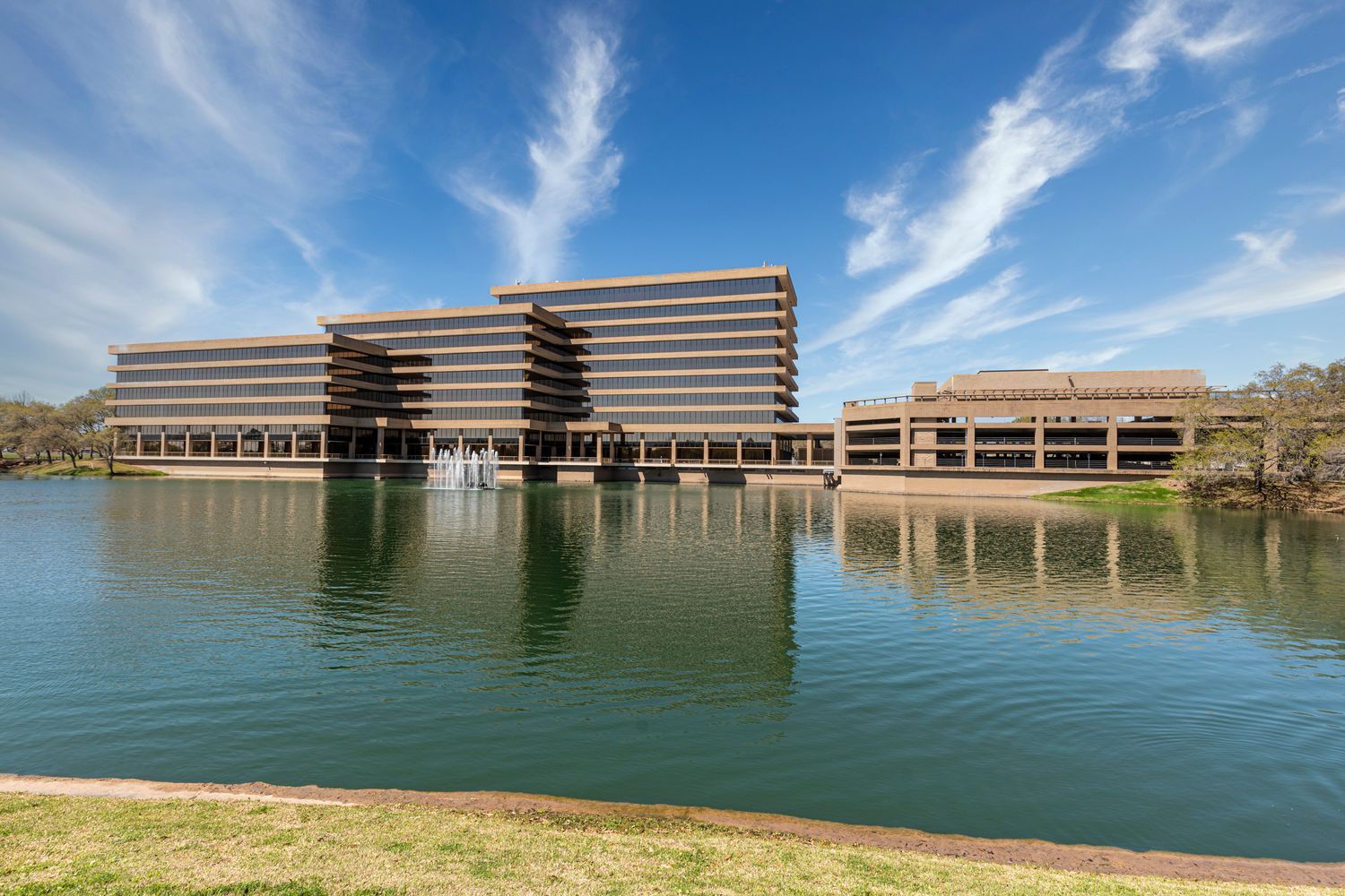 Modern, multi-story office building with a glass facade reflects in a large pond, set against a clear blue sky with wispy clouds.