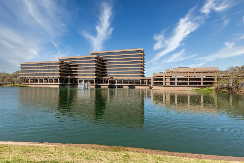 Modern, multi-story office building with a glass facade reflects in a large pond, set against a clear blue sky with wispy clouds.