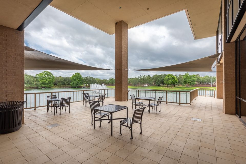 Outdoor patio with tables and chairs, overlooking a lake with a fountain and surrounded by trees, beneath a geometric canopy.