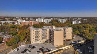 Aerial view of a cityscape with a large beige apartment building surrounded by trees and other buildings under a clear blue sky.
