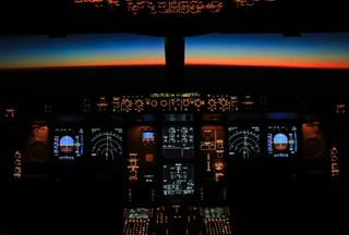 Aircraft cockpit with illuminated control panels, showing flight instruments against a vibrant sunset horizon in the background.