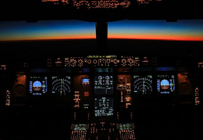 Aircraft cockpit with illuminated control panels, showing flight instruments against a vibrant sunset horizon in the background.