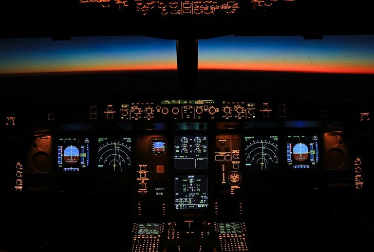 Aircraft cockpit with illuminated control panels, showing flight instruments against a vibrant sunset horizon in the background.