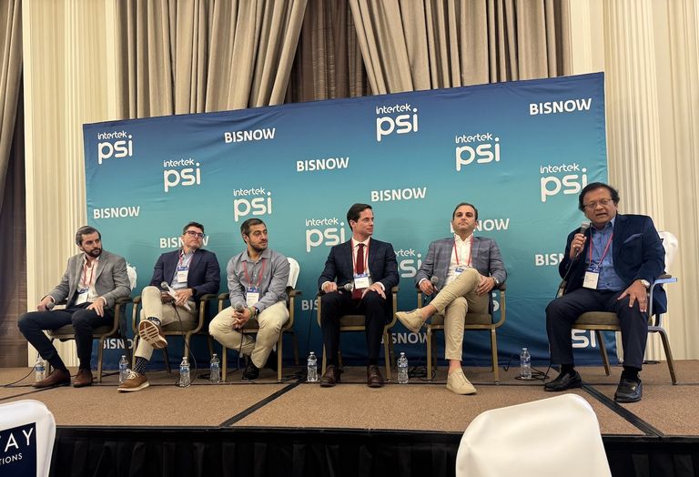 Six men sit on a panel stage discussing in front of a backdrop with "intertek PSI" and "BISNOW" logos.