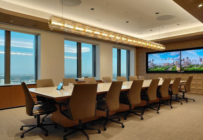 A modern conference room with a long table, leather chairs, large windows overlooking a city, and dual screens displaying a cityscape image.