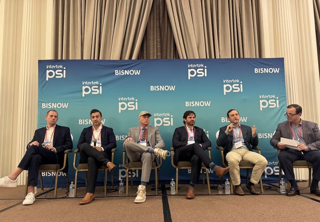 Six men sitting on a panel stage with a blue backdrop featuring logos of "Intertek PSI" and "BISNOW", engaged in a discussion.