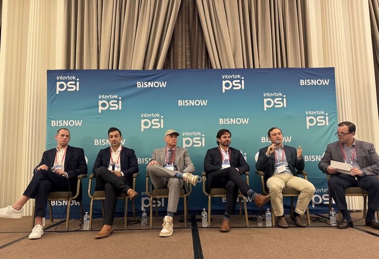 Six men sitting on a panel stage with a blue backdrop featuring logos of "Intertek PSI" and "BISNOW", engaged in a discussion.